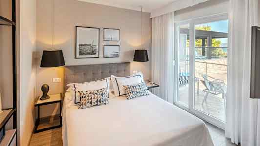 Modernt hotellrum med dubbelsäng, dekorerat med kuddar och konst på väggen. Glasdörrar leder ut till en möblerad terrass. (AI-genererad text)