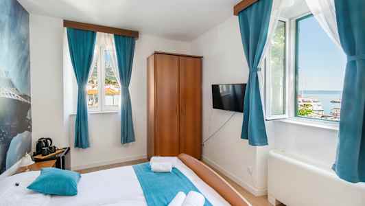 Hotellrum med dubbelsäng, blåa gardiner och utsikt över en hamn utanför fönstret. (AI-genererad text)