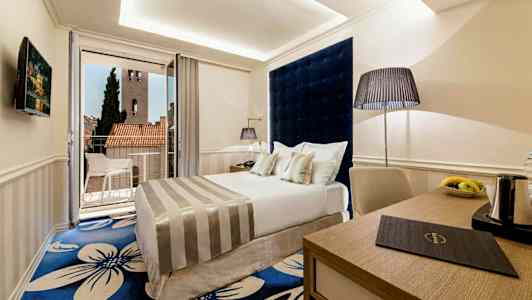 Ett hotellrum med en dubbelsäng, en fåtölj och ett skrivbord. Dörrar leder ut till en balkong med utsikt över takåsar och ett torn. (AI-genererad text)