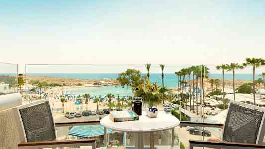 Vy från en balkong med bord och stolar mot en strand med parasoller och palmer vid havet. (AI-genererad text)