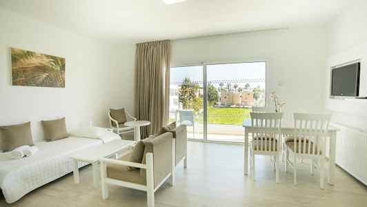 Ljust möblerat rum med säng, stolar, bord, och utsikt mot grönområde genom stora fönster. (AI-genererad text)