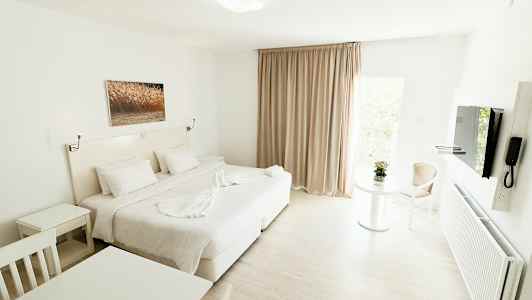Ett ljust och modernt hotellrum med en stor säng, vita möbler och en glastavla på väggen. Ett bord med en blomma och en stol syns vid ett fönster med beige gardiner. (AI-genererad text)