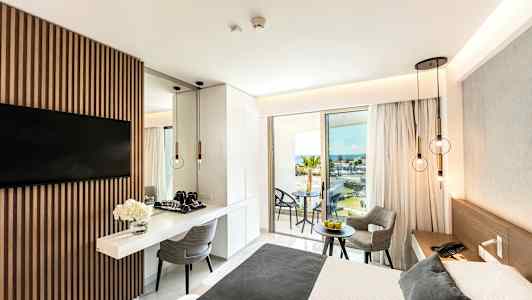 Modern hotellrum med en dubbelsäng, bord och stolar samt utsikt över havet från en balkong. (AI-genererad text)