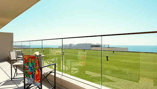 En terrass med glasräcke, stolar och en utsikt över gröna gräsmattor och havet. (AI-genererad text)