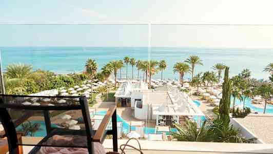 Utsikt över havet, hotellområde med palmträd och pooler, och en stol i förgrunden. (AI-genererad text)