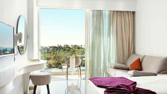 Ett ljust hotellrum med en soffa, en säng med lila täcke och en balkong som har utsikt över grönska. (AI-genererad text)