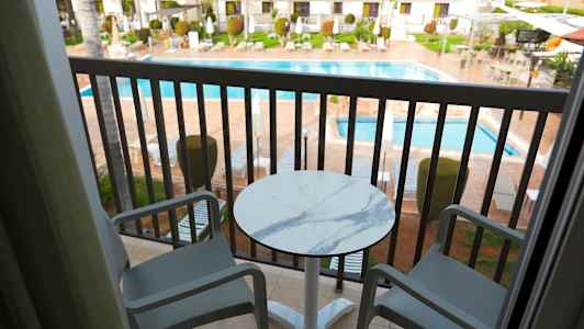 Två stolar och ett bord på en balkong med utsikt över en pool och omgivande byggnader. (AI-genererad text)