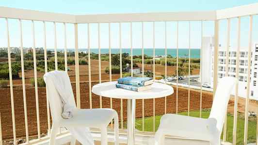 Två vita stolar och ett bord med böcker på en balkong med utsikt över jordbruksmark och havet. (AI-genererad text)