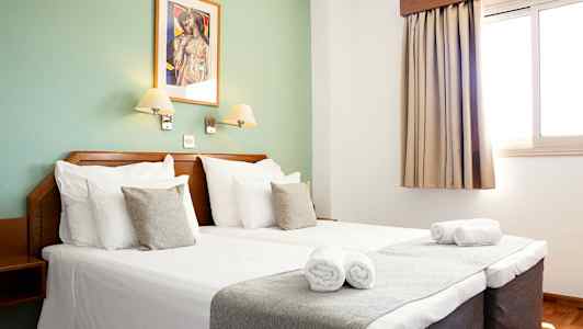 Interiör av ett hotellrum med dubbelsäng, två sänglampor, ett fönster med gardin och en dekorativ väggmålning. (AI-genererad text)