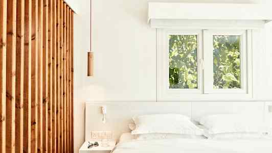 Ljus och luftig sovrumsinteriör med en säng, vita sängkläder och en träpanel samt fönster med grönskande utsikt. (AI-genererad text)