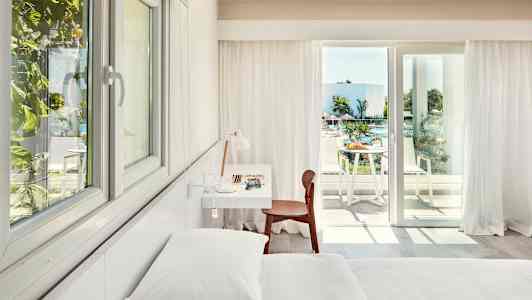 Interiör av ett ljust hotellrum med säng, skrivbord och stol, samt en balkong med utsikt över en pool och grönska. (AI-genererad text)