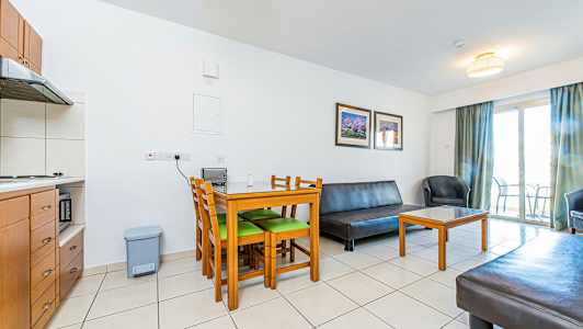 Ett vardagsrum med möbler inklusive ett bord med stolar, en soffa och en fåtölj. Ett kök syns i hörnet och en balkong med dörr i bakgrunden. (AI-genererad text)