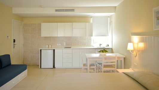 En ljus och modern studio med köksområde innehållande vita skåp, en litet bord med stolar och en sittmöbel mot en beige vägg. (AI-genererad text)