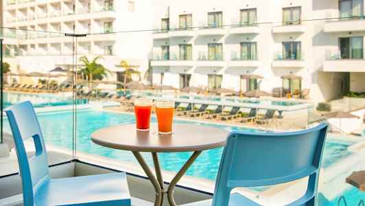 Två färgglada drycker står på ett bord vid sidan av två stolar, med en pool och hotell i bakgrunden. (AI-genererad text)