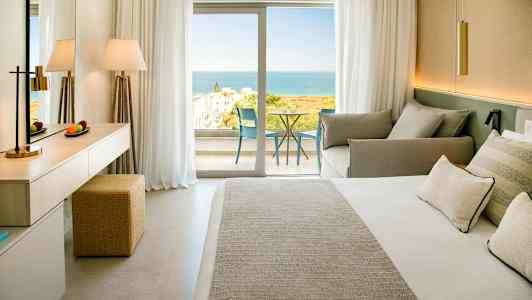 Ett hotellrum med en dubbelsäng, sittgrupp och en balkong med utsikt mot havet. (AI-genererad text)