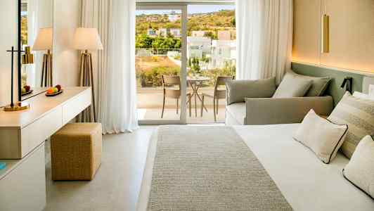 Bild av ett hotellrum med säng, soffa och skrivbord, samt en balkong med utsikt över ett bostadsområde. (AI-genererad text)