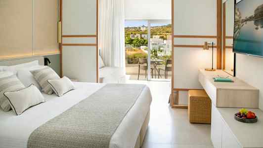 Ett ljust hotellrum med en stor säng, en arbetsyta, och en balkong med utsikt över grönska och byggnader. (AI-genererad text)