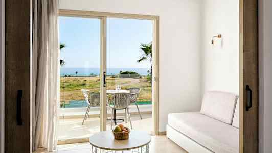 Ett rum med en soffa, ett bord och glasdörrar som leder ut till en terrass med bord och stolar, med havsutsikt. (AI-genererad text)