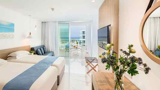 Ett hotellrum med två sängar, en soffa, ett skrivbord med blommor, stor spegel, och en balkong med utsikt mot havet. (AI-genererad text)