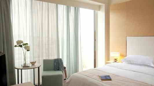 Ett sovrum med en bäddad säng, en stol, en bordslampa och ett fönster med ljusa gardiner. (AI-genererad text)