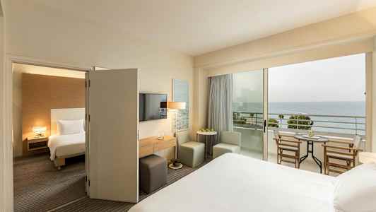 Ett hotellrum med en säng, sittplatser och ett bord, och en balkong med utsikt över havet. (AI-genererad text)