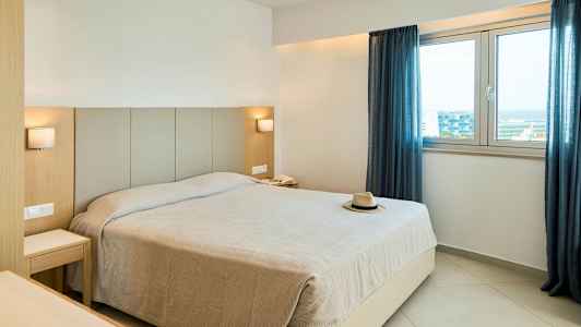 Ett hotellrum med en stor säng, två sängbord, två lampor, ett fönster med blå gardiner och en hatt på sängen. (AI-genererad text)