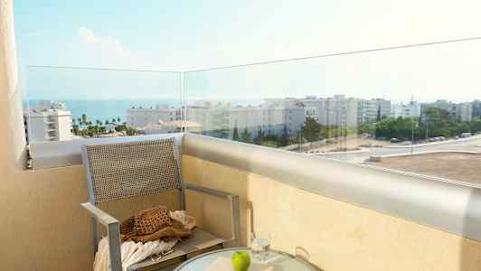 En balkong med utsikt över havet och byggnader, med en stol och ett bord med glas, bok och ett äpple. (AI-genererad text)