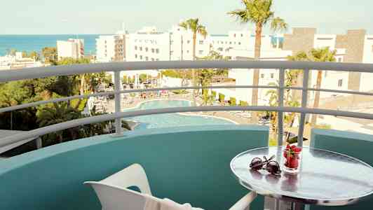 Utsikt från en balkong med utsikt över en pool och omgivande byggnader; ett litet bord har solglasögon och ett glas med jordgubbar. (AI-genererad text)