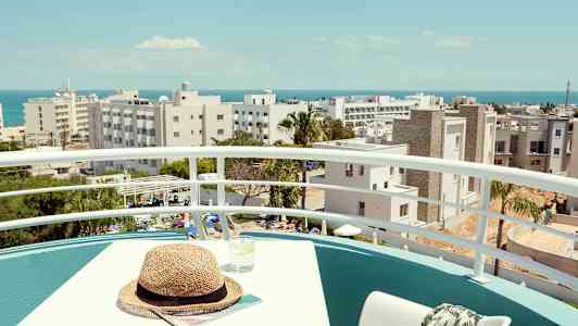 En balkong med bord, stol och utsikt över byggnader och havet i bakgrunden. (AI-genererad text)