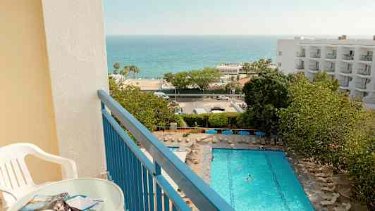 Utsikt från en balkong med bord och stol över en pool, grönska och havet. (AI-genererad text)