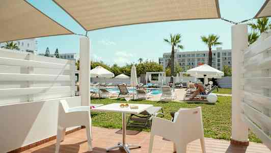 Utsikt från en terrass med vita stolar mot en pool omgiven av solstolar och parasoller. (AI-genererad text)