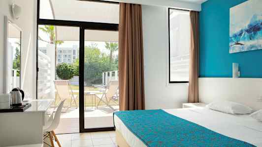 Modernt hotellrum med säng som har blått överkast, fönster öppnar mot terrass med stolar. (AI-genererad text)