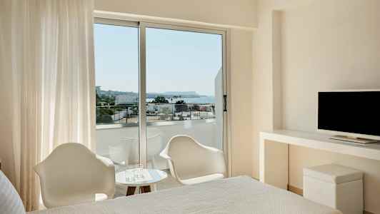 Ett hotellrum med en säng, ett skrivbord med en TV och stora fönster som öppnar upp till en balkong med utsikt över havet. (AI-genererad text)