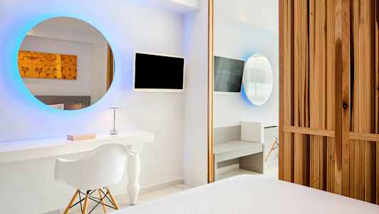 Ett modernt hotellrum med ett runt upplyst spegel, TV-skärm, ett skrivbord och en stol. Väggarna är vita och möblerna har minimalistisk stil. (AI-genererad text)