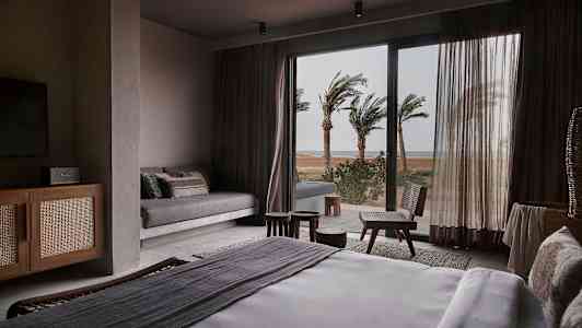 Ett hotellrum med en säng och utsikt över palmer utanför ett glasfönster. (AI-genererad text)