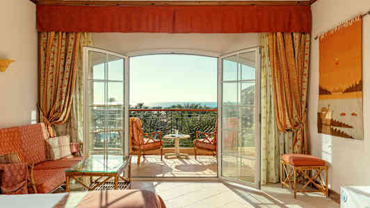 Ett hotellrum med öppen balkongdörr, sittmöbler och utsikt mot havet. (AI-genererad text)