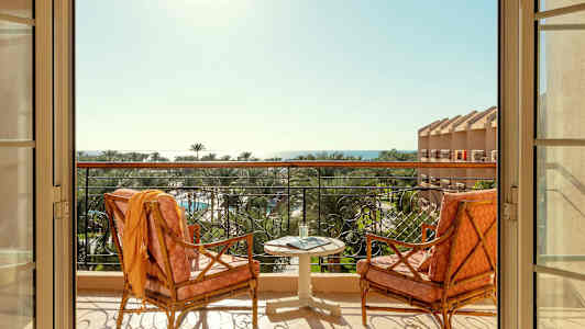 Två stolar och ett bord står på en balkong med utsikt över palmer och hav. (AI-genererad text)