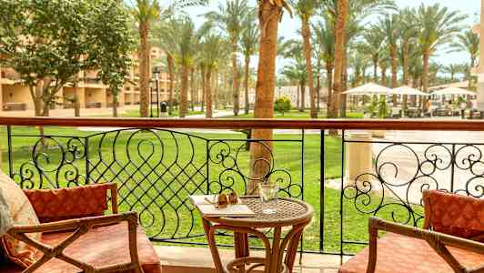 En balkong med stolar, ett bord med glas och solglasögon, och utsikt över en trädgård med palmer. (AI-genererad text)