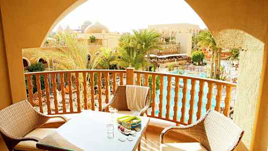 Balkong med möbler med utsikt över palmer, pool och sandfärgade byggnader. (AI-genererad text)