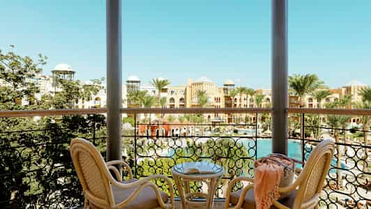 Utsikt från en balkong med två rottingstolar och ett bord. Bakom räcket syns palmer, en pool och byggnader med färgglada fasader. (AI-genererad text)