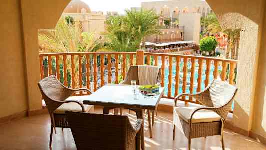 Vy från en balkong med bord och stolar samt utsikt över en pool och palmer vid en arkitektonisk byggnad. (AI-genererad text)