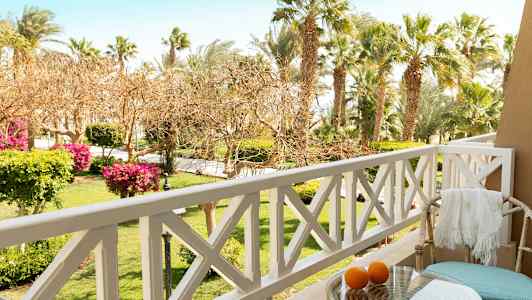 Utsikt från en balkong med utomhusstolar, bord och apelsiner, med en trädgård och palmer i bakgrunden. (AI-genererad text)