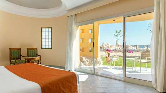 Rum med säng och bäddset i orange, två stolar med ett bord och utsikt mot en terrass med grönska och solstolar. (AI-genererad text)