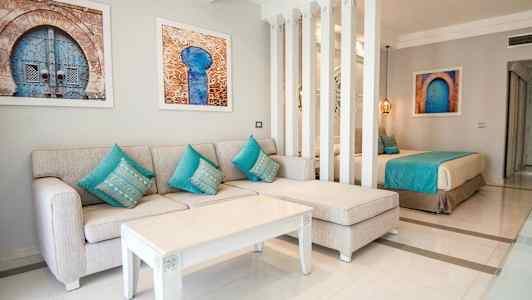 Ett vardagsrum med en beige soffa och turkosa kuddar, ett vitt soffbord och konstverk på väggen, delvis avskiljt med pelare från en sovdel med en säng. (AI-genererad text)