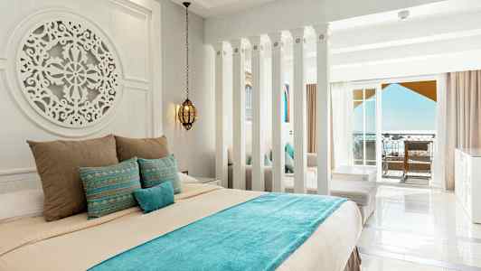 Ett hotellrum med ljusa väggar, en dekorerad säng med blå och beige sängkläder, samt en balkong med havsutsikt. (AI-genererad text)