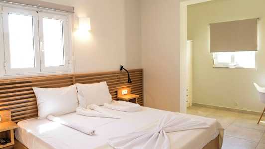 Ett hotellrum med en dubbelsäng, träpaneler som sänggavel, och en fönsterlampa ovanför. Ett litet bord med en läslampa och en öppning till en annan del av rummet. (AI-genererad text)