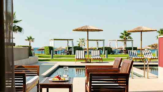 Utemöbler vid en pool med solstolar och parasoller, gräsmatta och hav i bakgrunden. (AI-genererad text)