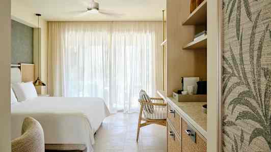 Ett hotellrum med en stor säng, en fåtölj och hyllor med kaffemaskin, belyst av naturligt ljus genom stora gardiner. (AI-genererad text)