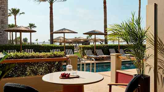 Uteplats med bord, stolar, utsikt över pool och solstolar under parasoller. (AI-genererad text)