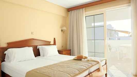 Ett hotellrum med en dubbelsäng, ljusa gardiner och en balkong med utemöbler. (AI-genererad text)